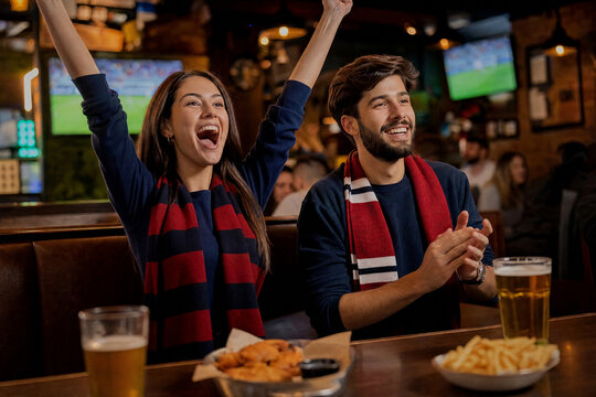 Excited young couple cheering for their favorite team while watching a soccer match in a sports bar with beer and snacks on the table during a weekend game night.