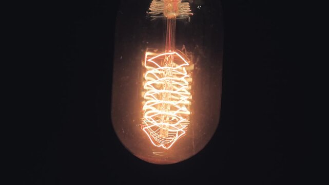 A close-up of an incandescent light bulb with a beautiful, unique filament hanging in the darkness and swinging before it goes out due to a power outage