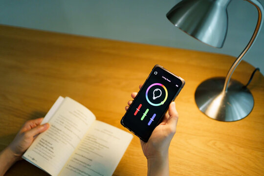 Hands holding smartphone with smart home lighting control app next to desk lamp and open book on wooden table in evening