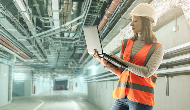 Construction engineering and infrastructure inspection. Female engineer checks technical systems with laptop in industrial utility corridor.