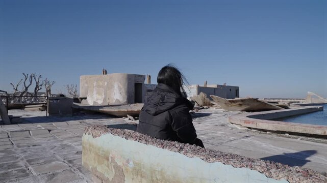 Woman seated in Villa Epecu&eacute;n Buenos Aires Argentina flooded town ruins with salt crust surfaces collapsed structures and dead vegetation showing post disaster environment, drone orbit shot