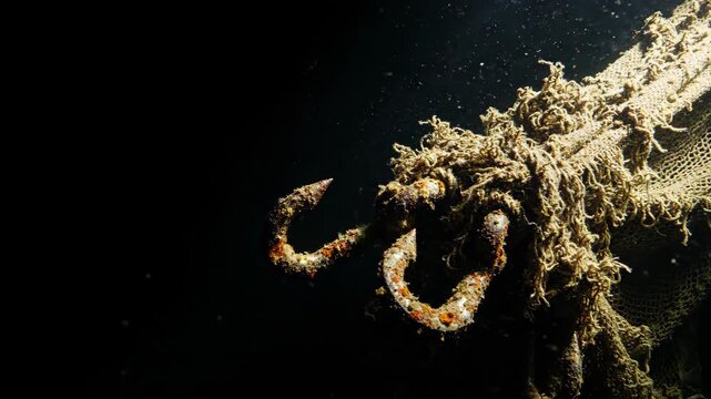 Rusty metal fishing hook attached to old net floating in deep dark water. Underwater shot of abandoned commercial fishing gear discarded in ocean habitat environment