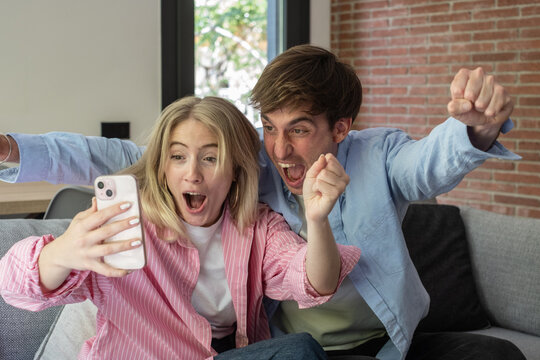 Excited Young Couple Cheering While Looking At Phone Together On Couch At Home