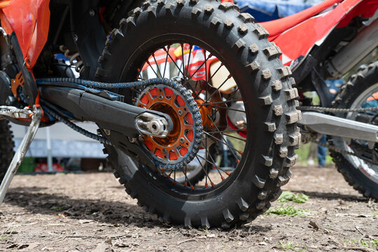 Close-up of a rear knobby tire, orange rear sprocket, and drive chain on an off-road motocross motorcycle outdoors. Detail view of a dirt bike rear wheel mechanism, drive chain