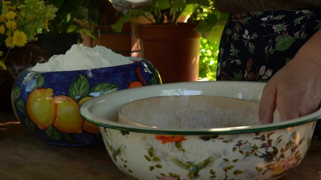 An elderly woman uses a wooden sieve to sift flour into a floral enamel bowl then adds salt sugar and water to mix a fresh batch of dough on a rustic wooden table in a garden setting
