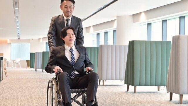 オフィスで車椅子利用者をサポートしながら移動するビジネスマンのバリアフリーの支援や障害者雇用などのイメージの動画