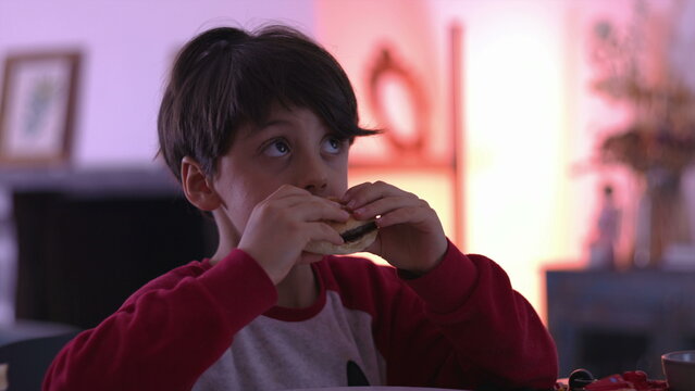 Young boy eating hamburger at dining table, taking big bite during indoor family mealtime and showing playful satisfaction and hearty appetite