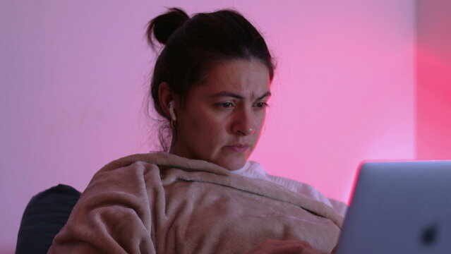 Close up of woman wrapped in blanket working on laptop at night, focused and serious while managing remote tasks and online communication from home