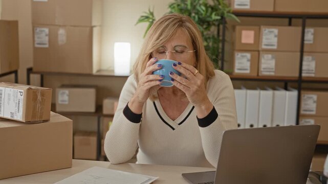 Woman holds mug with both hands amid stacked packages and laptop in a building office; quiet contentment.