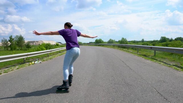 A grown woman in jeans learns to roller skate on an asphalt road.