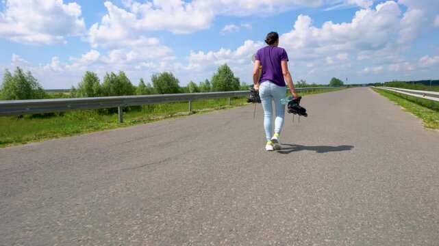 An adult woman in jeans walks along an asphalt road and carries roller skates.