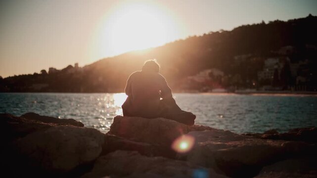 Lone man sitting on rocky beach shore looking at sun during peaceful sunset.