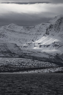 View of dramatic snow-covered mountains rising above a dark fjord under a moody, cloudy sky in Tromso, Troms, Norway.