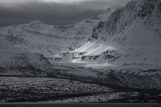 View of snow-covered mountain peaks under a dramatic cloudy sky with a beam of sunlight hitting the slopes Tromso, Troms, Norway.