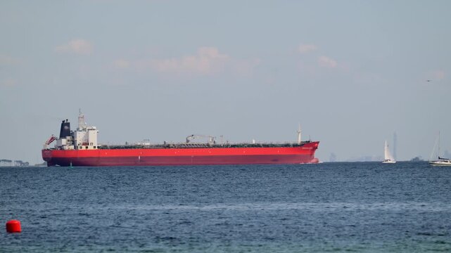 Large red oil tanker ship sailing across calm blue ocean water with sailboat in distance.