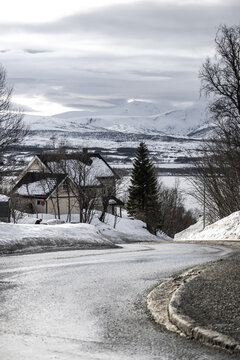 View of a winding icy road leading past snow-covered houses towards distant white mountains under a cloudy sky in Tromso, Troms, Norway.