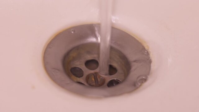 Close-up of a sink drain strainer with round holes as water flows through it. Focus on water movement and metal texture in a simple bathroom interior.