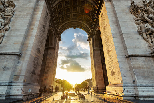 View of the Arc de Triomphe looking through the massive stone archway at sunset with people gathered at the base and intricate carvings on the walls Paris, Ile-de-France, France.