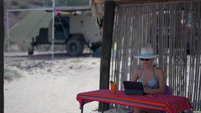 Mature woman working on computer tablet, and phone near camping teardrop trailer at tropical beach during vacation time in warm weather.