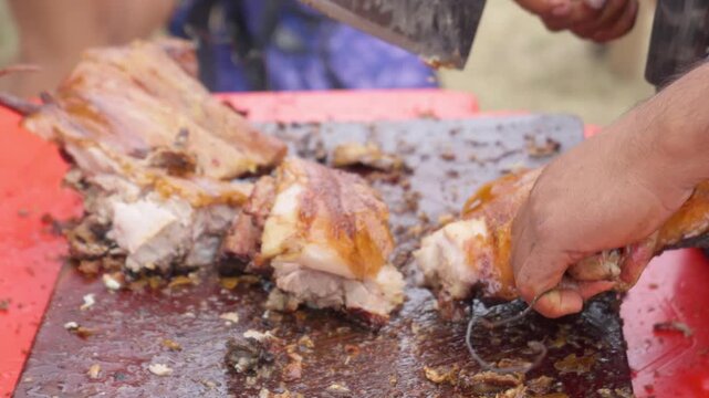 Mano cortando chancho a la cruz con hacha de carnicero sobre tabla de madera durante tradicional asado al fuego de le&ntilde;a sudamericano. 