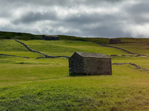 View of traditional stone barns and dry stone walls across the rolling green hills of the Yorkshire Dales under a cloudy sky Hawes, England, United Kingdom.