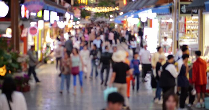 Blurred view of citizens strolling along wide pedestrian shopping street in downtown Kunming, passing brightly lit retail storefronts and marketplace stalls with neat civil twilight adding to scene.