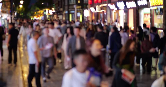 Defocused footage of anonymous people walking through busy pedestrian shopping street in downtown Kunming at night, glowing storefronts and neon signs creating vibrant bokeh effect