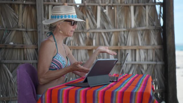 Mature woman shows emotion while using a computer, tablet, and phone at a tropical beach during her vacation time on a sunny day.