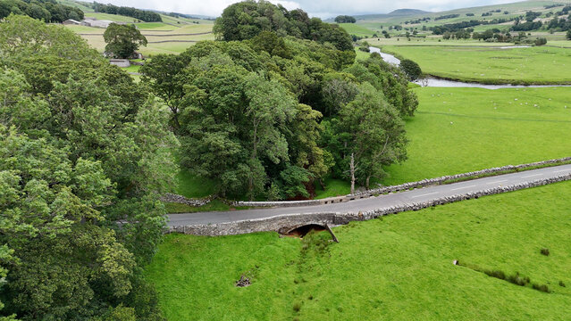 View of the Yorkshire landscape featuring a historic stone bridge over a stream surrounded by lush pastures and trees in Hawes, England, United Kingdom.