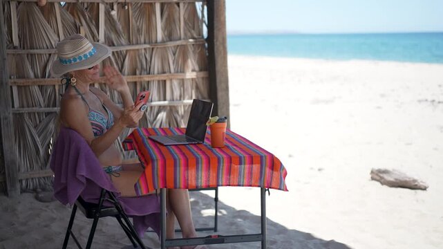 Mature woman thinks about her future while using computer and phone at beach during vacation, combining work and leisure time in sun.