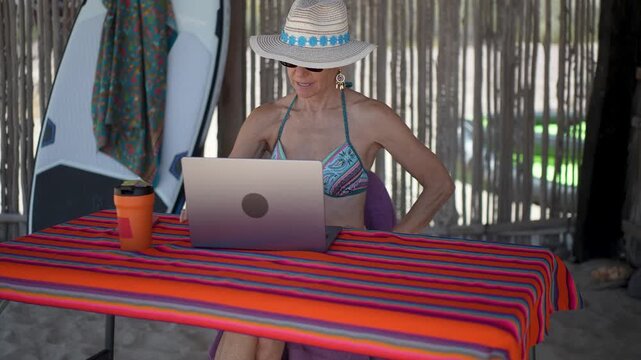 Mature woman engages in video chat on computer and phone at beach while experiencing different emotions during tropical vacation.