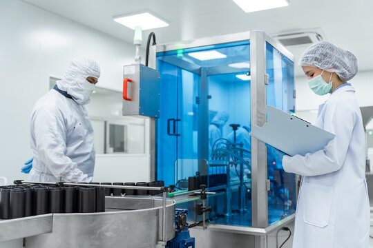 Clean room workers examine medical production machinery wit. Quality control inspector and factory workers operating automated packaging machinery in a sterile pharmaceutical manufacturing facility.