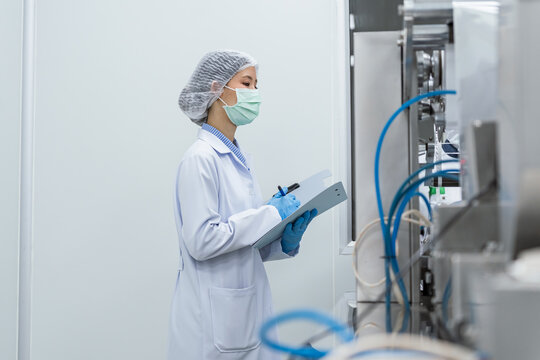 Medical technician reviewing data near factory machinery. Quality control inspector and factory workers operating automated packaging machinery in a sterile pharmaceutical manufacturing facility.