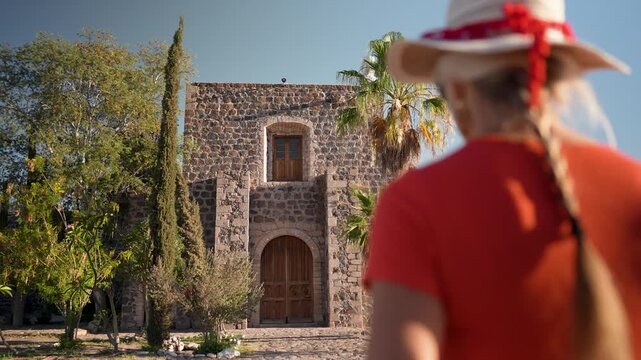Mature woman walks to Mission Church in Mulege, Baja California Sur, surrounded by palm trees and mountains.
