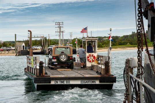 View of the Chappaquiddick ferry On Time III carrying a vehicle across the water with crew members and an American flag Edgartown, Massachusetts, United States.