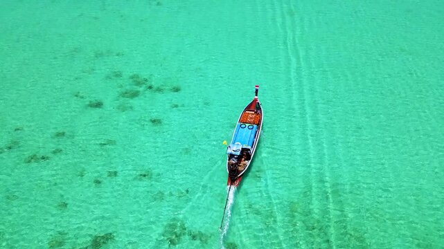 A turqouse colored ocean with clear water at Koh Kradan, a tropical island in Trang, Thailand, and a coral reef in front of a tropical island with a longtail boat, drone top view