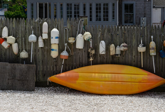 View of a yellow kayak resting on white gravel in front of a wooden fence decorated with weathered fishing buoys in Edgartown, Massachusetts, United States.