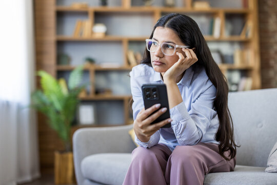 Young woman wearing eyeglasses and experiencing boredom or loneliness while sitting on a comfortable sofa at home, holding and looking distractedly at her smartphone during free time indoors
