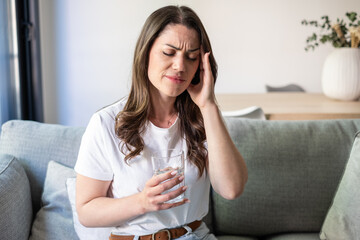 Obraz premium Young Woman Holding Glass Of Water On Sofa Rubbing Temple In Pain From Headache