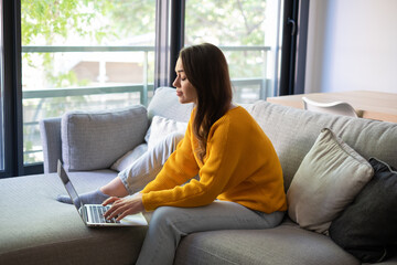 Fototapeta premium Young Woman Working On Laptop At Home On Couch In Bright Living Room