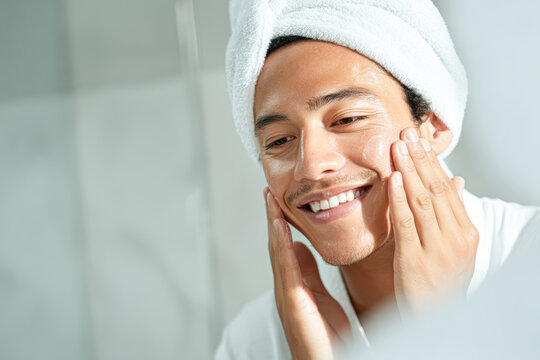 A man with a towel wrapped around his head applies skincare cream to his face, smiling in a bathroom.
