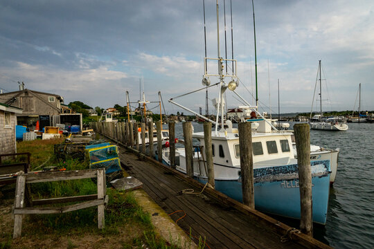 View of fishing boats docked at a wooden pier with lobster traps and fishing gear under a cloudy sky in Martha's Vineyard, Edgartown, Massachusetts, United States.