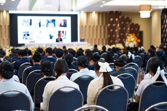 Large group of diverse students in uniforms sitting in a grand convention hall listening to an educational seminar with a speaker on stage, authentic academic conference and learning concept