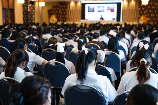 Large group of diverse students in uniforms sitting in a grand convention hall listening to an educational seminar with a speaker on stage, authentic academic conference and learning concept