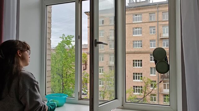 Young woman in rubber gloves cleaning window indoors while robot washer works on exterior glass; apartment building visible outside. Smart home meets traditional housekeeping.