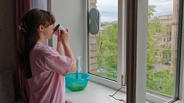 Window cleaning robot working on glass while woman relaxes with a cup of tea, watching the automated process. Smart home convenience, effortless housekeeping, modern lifestyle.