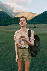 Fototapeta premium Young woman hiking in a mountain meadow holding a phone, backpack on her shoulder, wearing khaki shorts and a beige shirt.