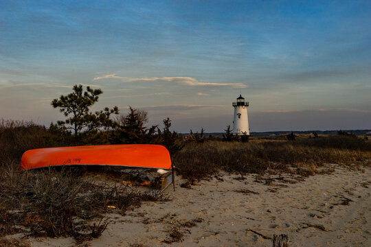 View of the Edgartown Harbor Light and an orange canoe overturned on a sandy beach with coastal vegetation under a twilight sky in Edgartown, Massachusetts, United States.
