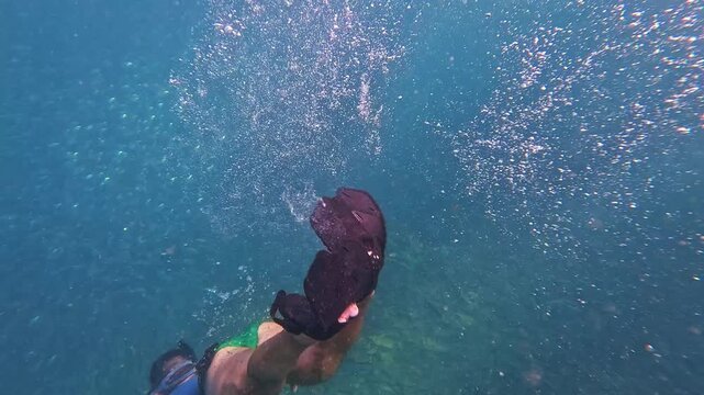 A person in a blue shirt and swim fins swims gracefully through clear ocean water, surrounded by an enormous school of small fish above a coral reef