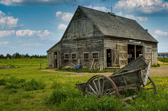 View of a rustic weathered wooden barn and an old cart in a green field under a cloudy sky in Edgartown, Massachusetts, United States.